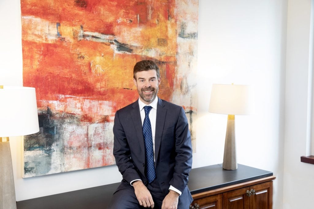 A man in a suit sits on a dark cabinet in front of a large abstract orange and white painting, with two table lamps on either side.