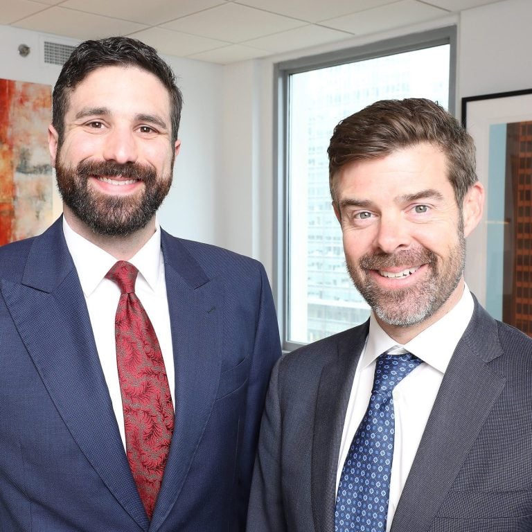Two men in suits stand indoors in a modern office setting, both smiling at the camera. Large windows and abstract artwork are visible in the background.
