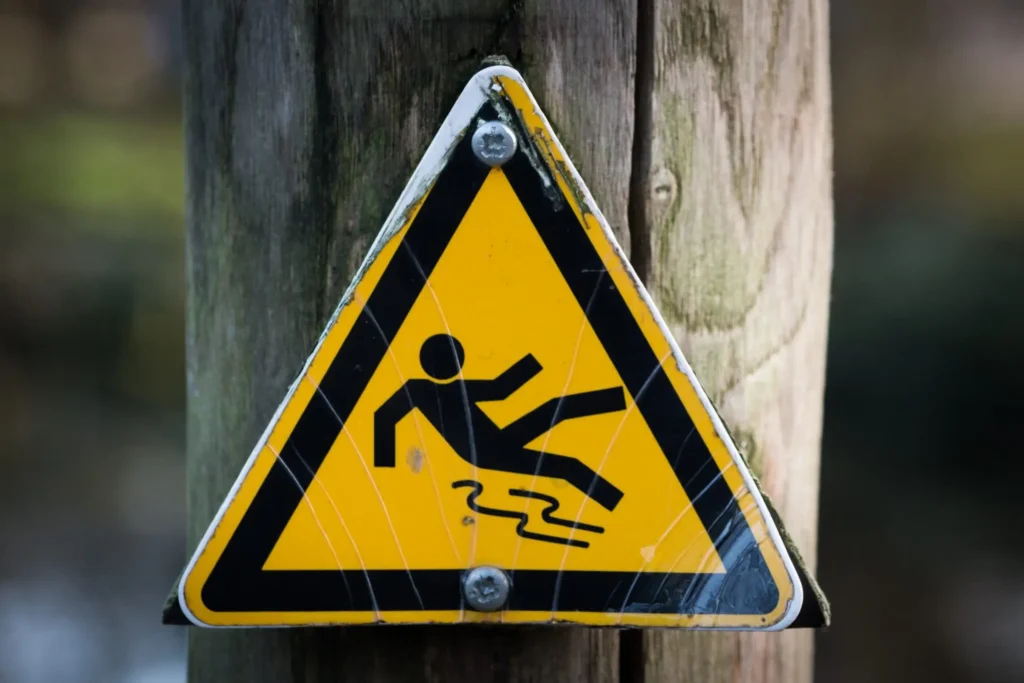 Yellow triangular warning sign attached to a wooden post, showing a person slipping to indicate a slippery surface.