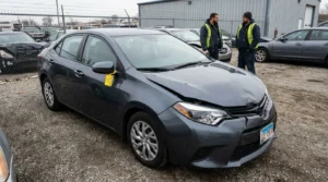damaged sedan with tagged door handle inspected by two workers at salvage yard