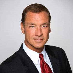 A man in a dark suit, white shirt, and red tie poses against a plain light background, looking directly at the camera with a neutral expression.